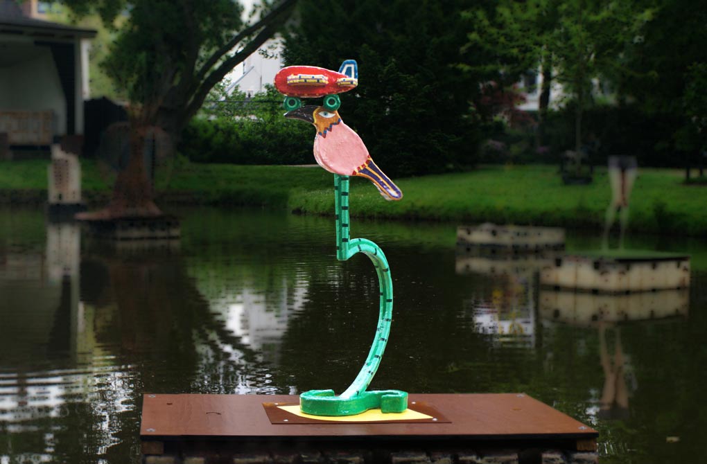 De titel is Voor de spelende mens, op de letter s is een verticaal gericht hefboomsleutel geplaatst. Op de deze sleutel zit een vogel. Op de kop van de vogel landt een vliegtuig. Het is 120 cm hoog, kleurrijk en gemaakt van polyster