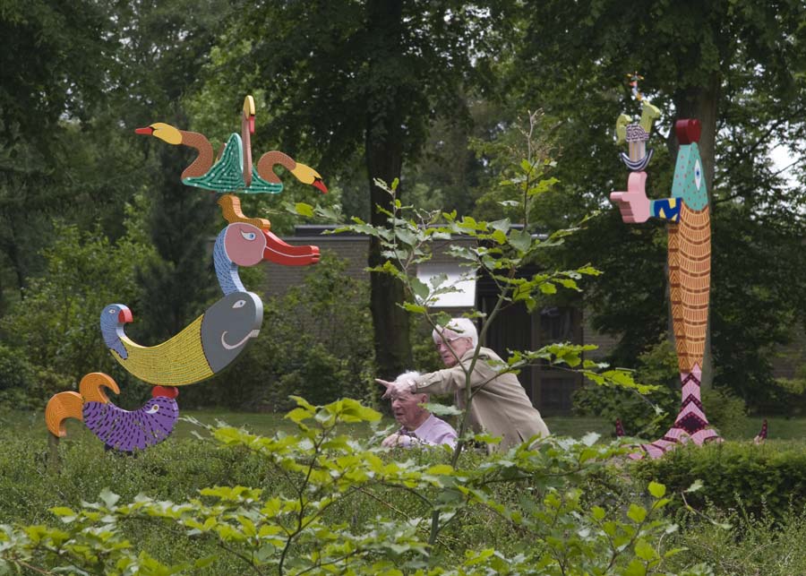 twee kinetische beelden op de tentoonstelling in Auxiliatrix, Venlo. Het beeld op de voorgrond is in veel kleuren beschilderd en beweegt op de wind. Van onder naar boven, een bewegende vis, op de bek van de vis draait een groot kuifeend. Op de kuif van de eend draaien vier zwanen.