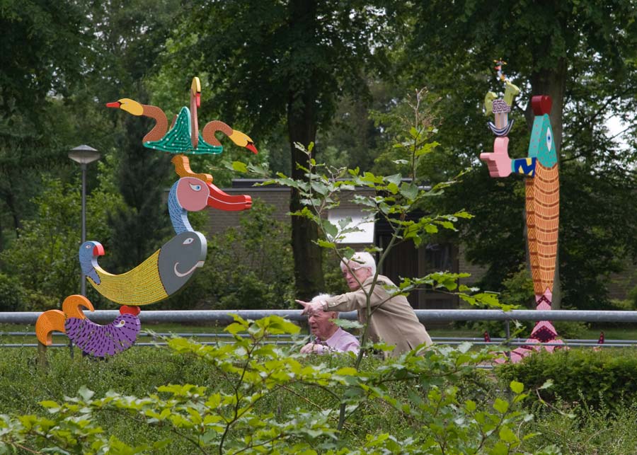 twee kinetische beelden op de tentoonstelling in Auxiliatrix, Venlo. Het beeld op de voorgrond is in veel kleuren beschilderd en beweegt op de wind. Van onder naar boven, een bewegende vis, op de bek van de vis draait een groot kuifeend. Op de kuif van de eend draaien vier zwanen.